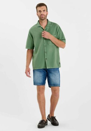 Kurzärmeliges Hemd zum Zuknöpfen aus hellgrünem Stoff mit Brusttasche, kombiniert mit blauen Jeansshorts und dunklen Schnürschuhen.
