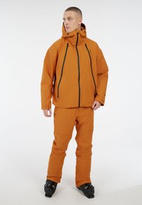 Narančasta vodootporna skijaška jakna s crnim patentnim zatvaračima, podesivim kapuljačom i suknjom u odgovarajućoj narančastoj boji. Teksturirani materijal, prilagođeni dizajn, nosi se s skijaškim čizmama.