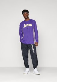 Chemise à manches longues violette avec logo "LAKERS" et accents blancs, associée à un pantalon de survêtement noir avec branding blanc, portée avec des baskets blanches.