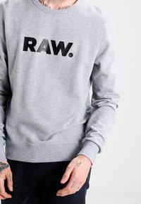 Sudadera gris con cuello redondo y puños acanalados, que presenta un gran texto negro "RAW." en la parte frontal. Material de algodón, textura suave.