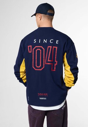 Mann mit dem Rücken zum Betrachter trägt ein marineblau-gelbes Langarmshirt mit "'04 Since" in rotem und weißem Text sowie dem Red Bull Racing-Logo auf dem Rücken.