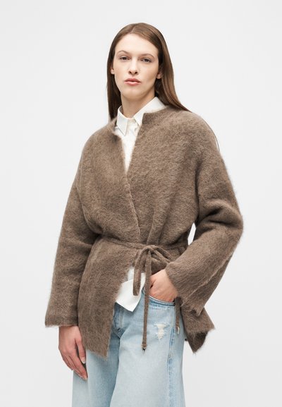 Γυναίκα που φοράει ένα μαλακό καφέ ζακέτα με ζώνη πάνω από ένα λευκό πουκάμισο και ανοιχτόχρωμα τζιν με σκισίματα, που στέκεται μπροστά σε μια απλή λευκή φόντο.