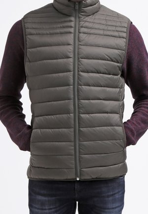 Hombre con chaleco acolchado y sin mangas gris sobre camisa de manga larga burdeos, con las manos en los bolsillos, mostrado desde el cuello hasta las caderas.