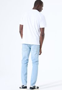 Camiseta blanca de algodón con mangas cortas, combinada con jeans de color azul claro con bolsillos traseros, llevada con zapatillas de deporte en blanco y negro.