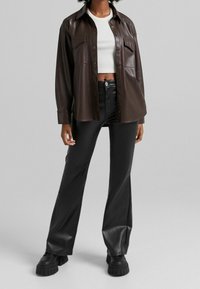 Brun konstläder skjortjaket med framfickor, bärs över en vit kort topp och svarta utsvängda läderbyxor, i kombination med kraftiga svarta stövlar.