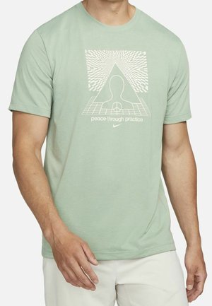 Homme portant un t-shirt vert clair avec un graphisme blanc d'un contour de tête, un triangle, et le texte "peace through practice" sur le devant.