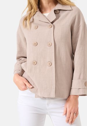 Frau trägt eine beige, doppelt geknöpfte Jacke mit langen Ärmeln und weiße Hose, Hand in der Tasche.