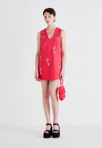 Red cotton shift dress with floral embroidery. Features a V-neck and sleeveless design. Accompanied by a small textured red handbag.
