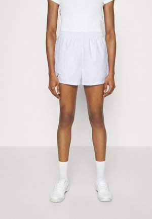 Person iført hvide atletikshorts, hvide crew-sokker, hvide sneakers og en hvid tætsiddende kortærmet skjorte, stående mod en ensfarvet væg.