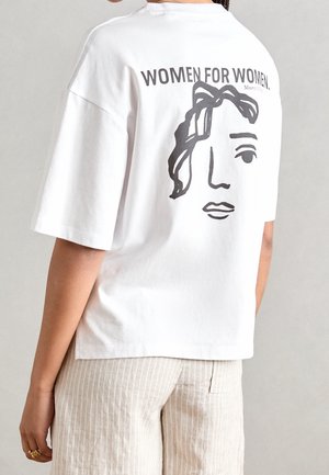 Henkilö, joka pukeutunut valkoiseen T-paitaan, jossa on musta abstrakti naisen kasvokuvio ja teksti "WOMEN FOR WOMEN" takana, yhdistettynä vaaleisiin raidallisiin housuihin.