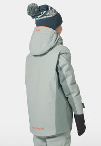 Světlé šedé voděodolné sako s kapucí, kontrastními černými rukávy a oranžovým logem. V páru s modrou pletenou čepicí s vzorem.