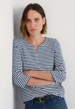 Haut à rayures bleu marine et blanc à manches longues, avec un col rond et des manches trois-quarts, associé à un jeans bleu foncé et une ceinture noire.
