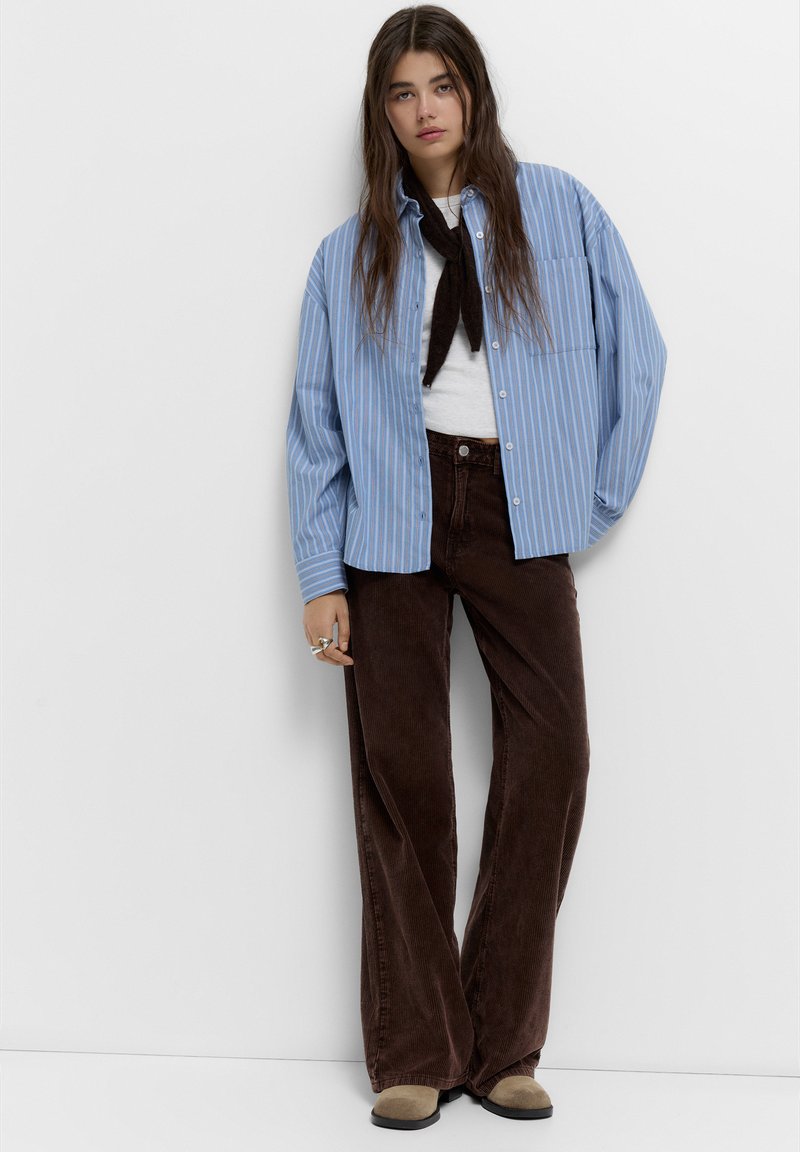 Camicia a righe blu e bianche con bottoni sopra una maglietta bianca, abbinata a pantaloni a gamba larga in velluto a coste marroni e scarpe beige. Gli accessori includono una sciarpa nera.