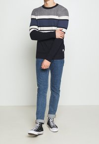 Man i randig marinblå, grå och vit tröja med uppvikta blå jeans och svarta högstövelsnickers som står mot en slät vägg.