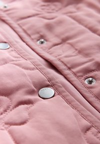 Closeup af pink quiltet stof med synlige sølv trykknapper, en af dem indgraveret med ordet "NEXT."