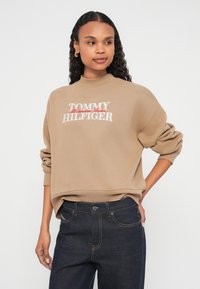 Καφέ φούτερ με υψηλό λαιμό και ριπ μανσέτες. Διαθέτει λευκό και κόκκινο κεντητό κείμενο "TOMMY HILFIGER" στο εμπρόσθιο μέρος. Εμφανίζονται σκούρα τζιν παντελόνια.