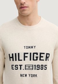 Maglione beige lavorato a maglia con scollo rotondo, caratterizzato da testo nero: "TOMMY HILFIGER EST. 1985 NEW YORK" ben visibile sul davanti.