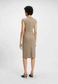 Form-fitting beige dress with cap sleeves, back zipper, and a rear vent, paired with black heeled slingback shoes. Smooth fabric texture.