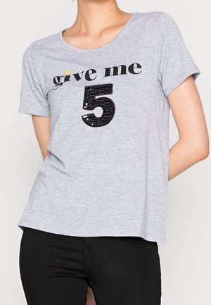 Hellgraues Kurzarmhemd mit dem Text "give me" und einer großen schwarzen Pailletten-Zahl 5, getragen von einer Person mit verschränkten Händen hinter dem Rücken.