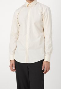 Camicia button-up color crema chiaro con maniche lunghe, colletto a punta e una texture sottile, abbinata a pantaloni neri.