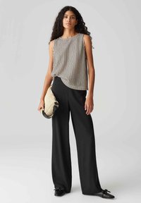 Schwarze weit geschnittene Hose, gestreifter ärmelloser Oberteil in hellen und dunklen Tönen sowie eine strukturierte Strohtasche, kombiniert mit schwarzen Schuhen.