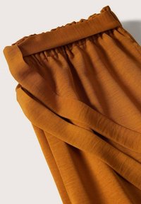 Pantalon en tissu orange rouille avec une taille élastique froncée et de larges plis aux jambes, présenté sur un fond clair uni.