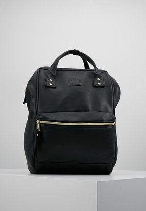 Sac à dos en cuir noir avec poignées supérieures et poche zippée à l'avant, posé sur un socle blanc sur un fond gris.