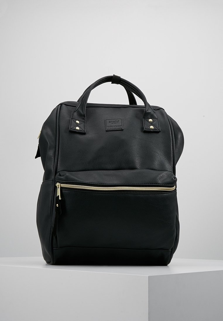 Sac à dos en cuir noir avec poignées supérieures et poche zippée à l'avant, posé sur un socle blanc sur un fond gris.