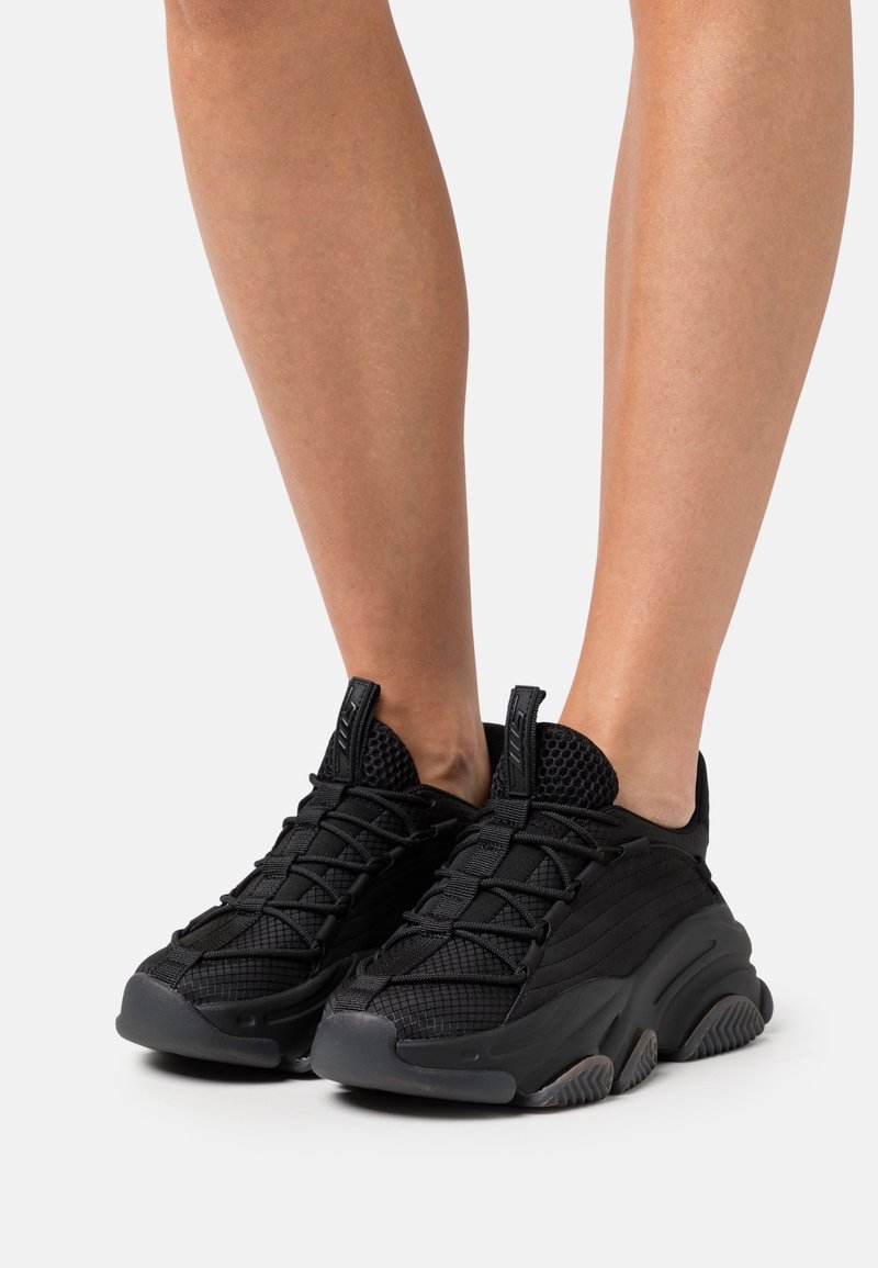 Des chaussures de sport noires avec une tige en mesh et des détails texturés, dotées d'une semelle épaisse en caoutchouc et d'un design épuré.