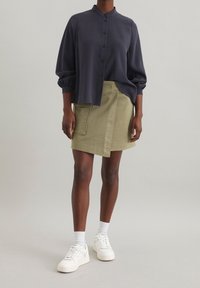 Marineblaues Hemd mit hohem Kragen und lockerer Passform, kombiniert mit einem khakifarbenen Wickelrock mit aufgesetzter Tasche, getragen mit weißen Sneakers.