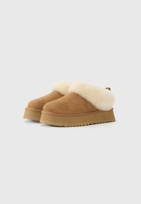 Scarpe slip-on in suede marrone con fodera spessa in pelliccia beige attorno al colletto e suole in gomma leggermente elevate su sfondo bianco.