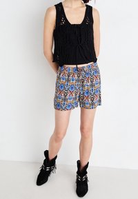 Top corto nero all'uncinetto con nodo frontale, abbinato a pantaloncini colorati con motivi floreali blu e rossi. Stivaletti neri alla caviglia.