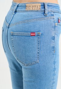 Gaiši zilos džinsa skinny džinsos ar vidēju vidukli, ar ādas logotipa uzlīmi, sarkano birku uz kabatas un minimālistiskiem dūrienu detalizējumiem.