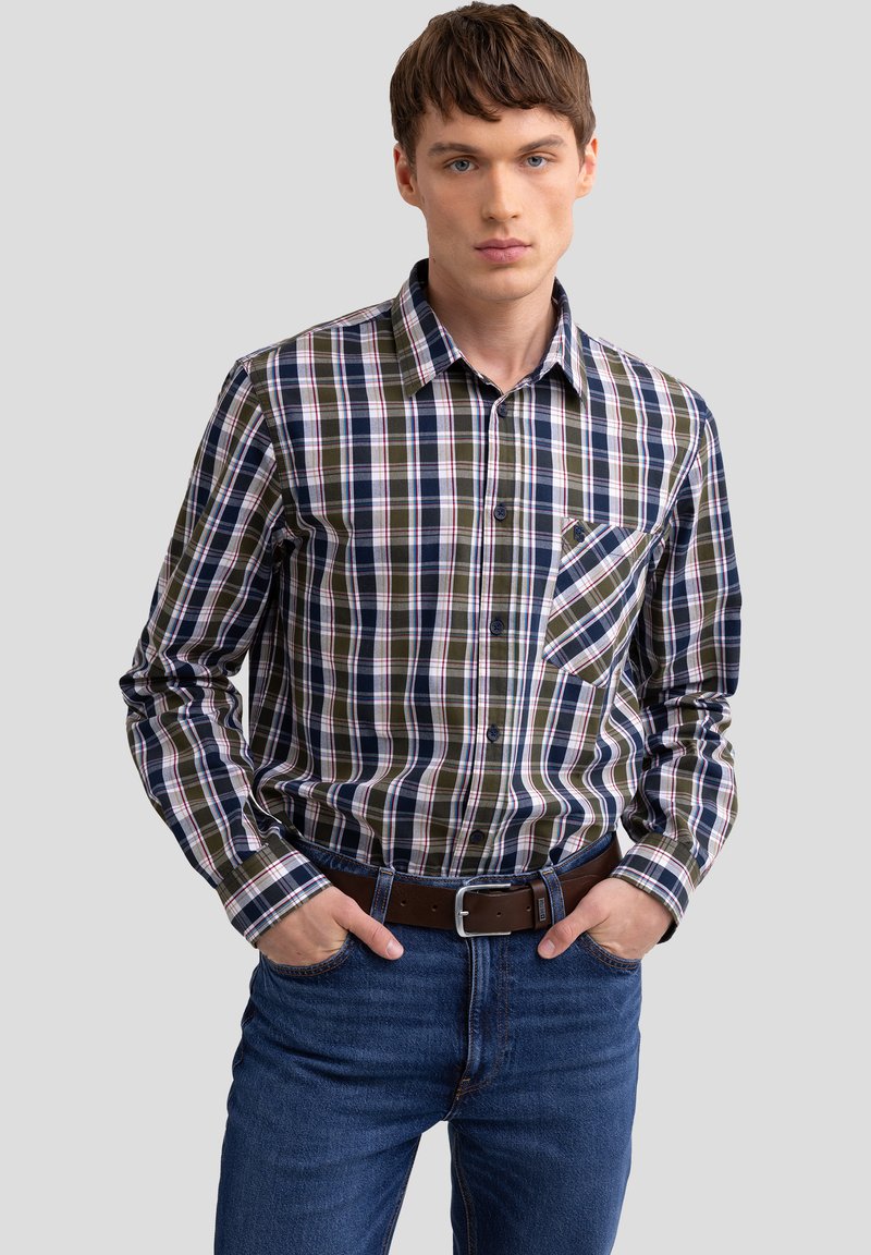 Camisa de cuadros de manga larga en tonos azul marino, verde y blanco, con cuello abotonado y bolsillo frontal. Combinada con jeans azules y un cinturón marrón.