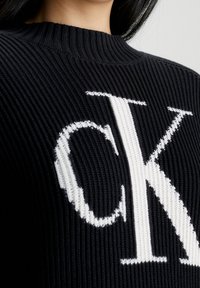 Sweatshirt en tricot noir à côtes avec un grand logo blanc 'CK' sur le devant. L'encolure est ronde et texturée, avec une silhouette ajustée.