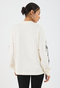 Long-sleeve cream shirt in soft fabric, featuring black graphic design on sleeves, relaxed fit, and ribbed cuffs.