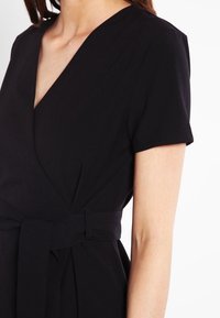 Musta kietaisumekko, jossa on V-pääntie, lyhyet hihat ja vyötärösolki. Sileä kangas, joka myötäilee vartalon muotoja ja siinä on minimaalinen ommeltyö.