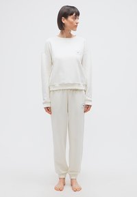 Γυναίκα που στέκεται ξυπόλυτη, φοράει ταίριαστο κρεμ φούτερ και φόρμα με μικρά λογότυπα, κοιτάζοντας προς τα δεξιά της μπροστά σε απλό φόντο.