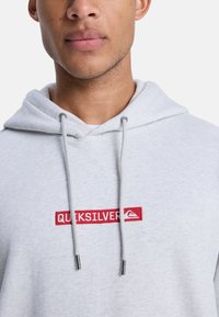 Sudadera gris claro con un logo bordado en rojo "QUICKSILVER" en el frente, cierre con cordón y una textura suave. Diseñada para la comodidad y el uso casual.