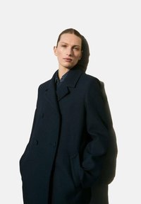 Donker marineblauwe dubbelbreasted wollen coat met geknipte kraag, zijzakken en een iets oversized pasvorm, gedragen over een blauwe blouse.