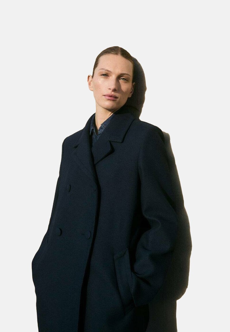Donker marineblauwe dubbelbreasted wollen coat met geknipte kraag, zijzakken en een iets oversized pasvorm, gedragen over een blauwe blouse.