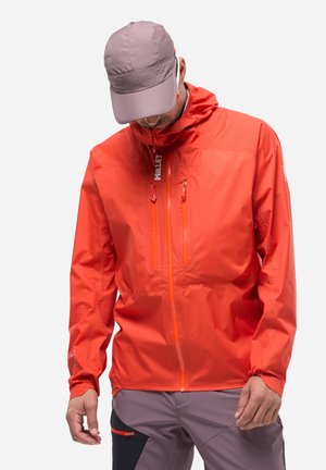 Homme portant une veste imperméable orange vif avec des fermetures éclair frontales et une casquette mauve, regardant vers le bas sur un fond blanc.