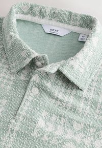 Grøn- og hvidmønstret polo skjorte med tekstureret stof. Har en button-down krave og to knapper ved halsen. Størrelser markeret.