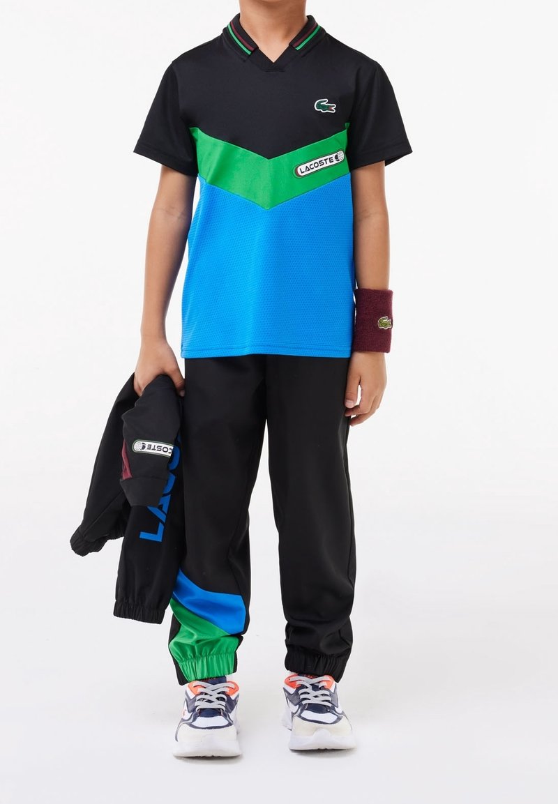 Black short-sleeve polo with green and blue chevron design, paired with black track pants featuring green and blue accents, and sneakers.
