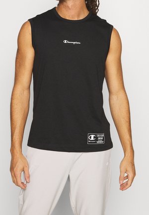 Mężczyzna ubrany w czarną, bezrękawnikową koszulkę Champion z logo na piersi i metką przy krawędzi, w połączeniu z jasnymi spodniami.