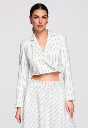 Žena s tmavými vlasy sepnutými do nízkého culíku, oblečená v bílém krátkém blejzru s proužky a odpovídající vysokým pasem sukně na jednoduchém pozadí.