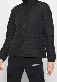 Svart puffjacka med hög krage, vertikal och diagonal quiltad design, med fickor med dragkedja och varumärkeslogotyp på vänster sida.