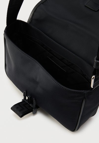 Μαύρη τσάντα ταχυδρόμου από νάιλον με λεία επιφάνεια, ρυθμιζόμενο λουράκι και κλείσιμο με πόρπη μπροστά. Το εσωτερικό διαθέτει πολλαπλά διαμερίσματα.