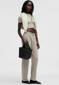 Beige Cropped-Top und passende Hose, mit einem cremefarbenen gestrickten Weste über den Schultern. Schwarze gesteppte Tragetasche in der Hand. Flache Schuhe.