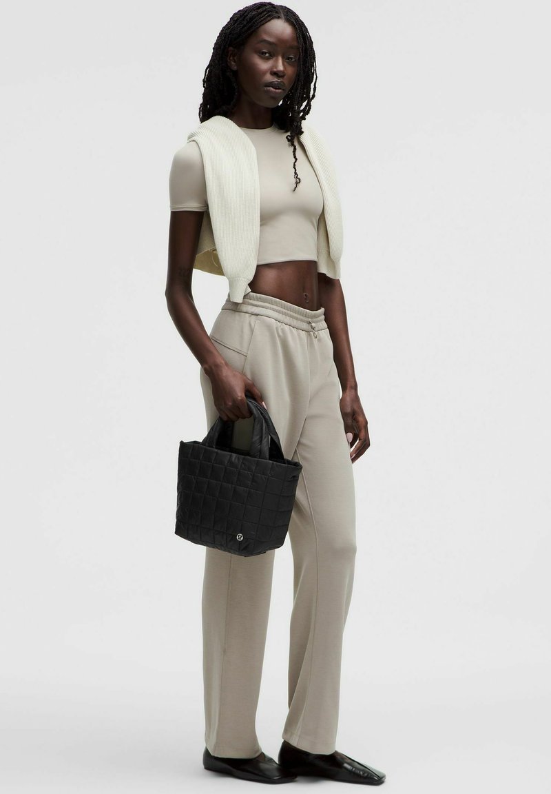 Beige Cropped-Top und passende Hose, mit einem cremefarbenen gestrickten Weste über den Schultern. Schwarze gesteppte Tragetasche in der Hand. Flache Schuhe.